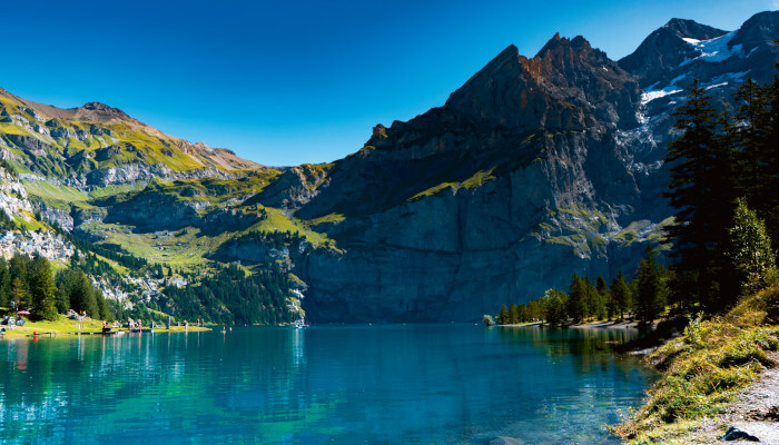 Blauer See in saftiggrünen Wiesen vor Felswand mit Bäumen